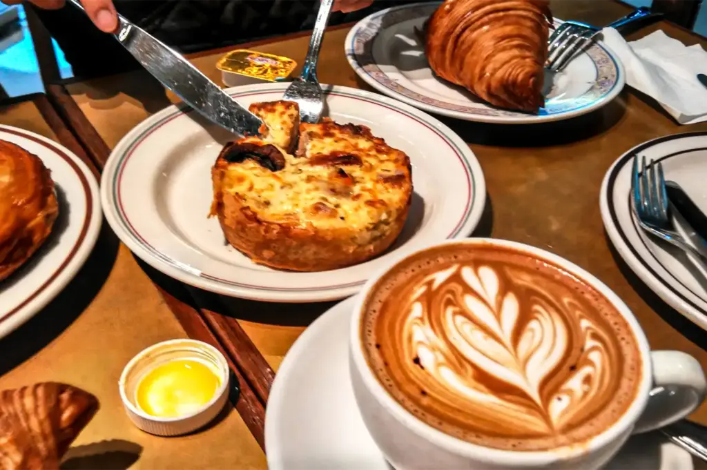 A person uses a fork and knife to slice into a deep-dish quiche on a ceramic plate at a cafe table. Surrounding the dish are butter packets, fresh croissants, and a cup of hot cappuccino detailed with elegant latte art foam.