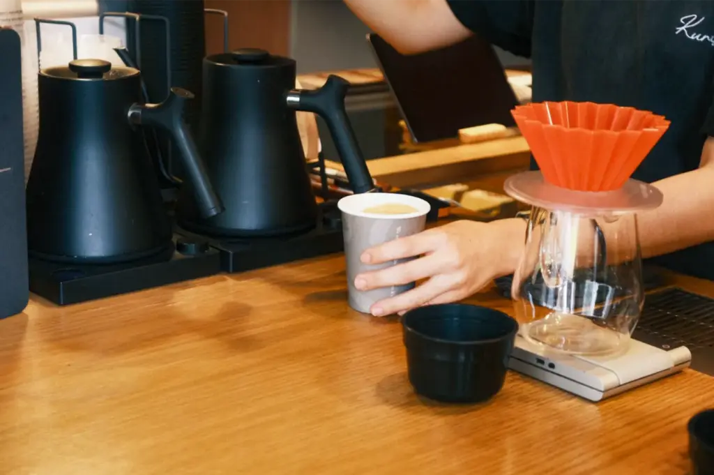 A barista places a freshly prepared takeaway coffee cup onto a warm wooden service counter lined with professional brewing equipment. Beside two matte black electric gooseneck kettles, an origami-style orange pour-over dripper rests atop a clear glass server on a precision digital scale.